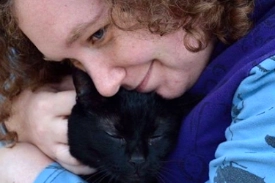 A girl with curly brown hair is hugging her black cat as she sits on the sidewalk because she found him after he had been missing.