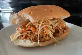 A pulled soy curls sandwich topped with coleslaw on an Italian bun sits on a white plate. The plant-based version of pulled pork.