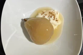 A white bowl with a single, whole poached pear accompanied by a dollop of yogurt topped with cinnamon.