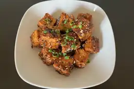 A white plate with sticky tofu puffs garnished with black and white sesame seeds and green onion.